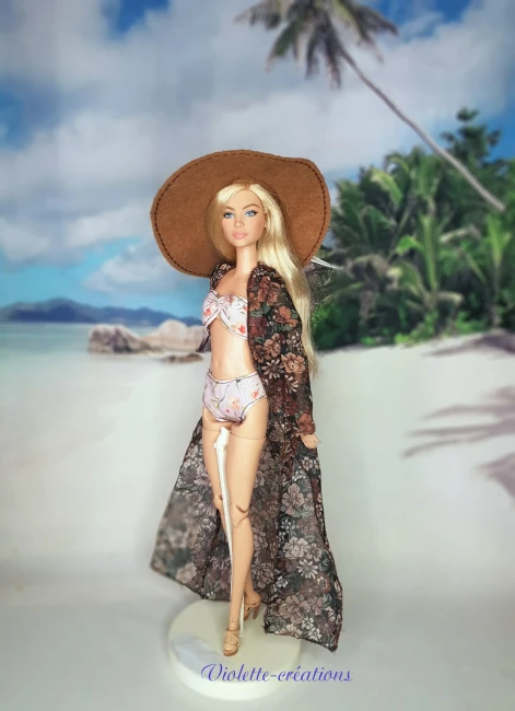 Poupée type Barbie portant un maillot de bain deux pièces pastel avec kimono fleuri transparent, chapeau de soleil et sandales sur un décor de plage grecque.