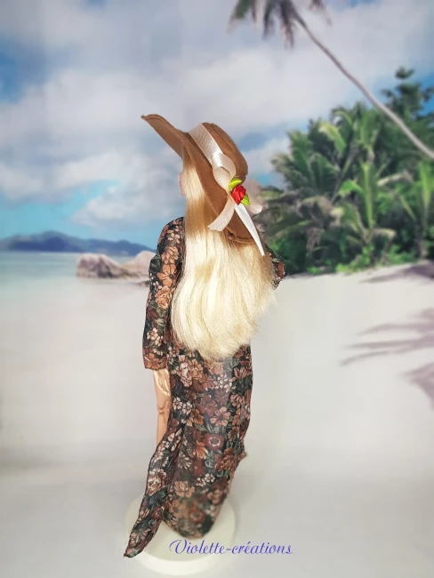 Poupée type Barbie portant un maillot de bain deux pièces pastel avec kimono fleuri transparent, chapeau de soleil et sandales sur un décor de plage grecque.