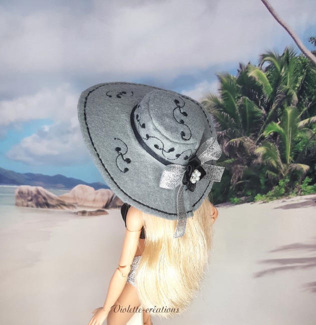 Maillot de bain 2 pièces pour poupée mannequin type Barbie avec haut noir, bas argenté et chapeau en feutrine sur décor de plage.