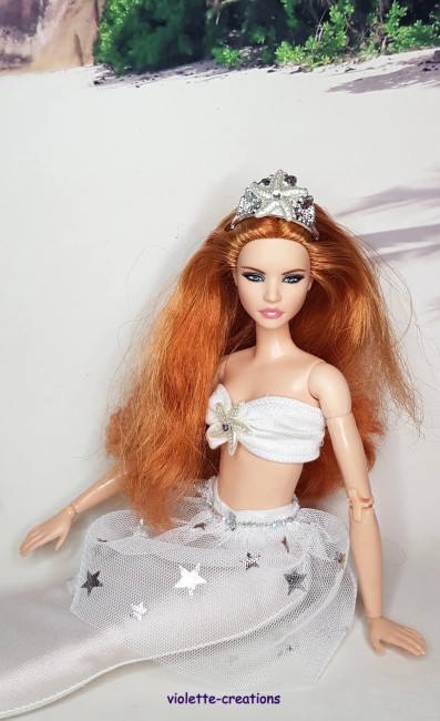 Barbie portant une tenue de sirène blanche scintillante avec effet strass et un haut assorti en forme de coquillage, posé sur un décor de plage