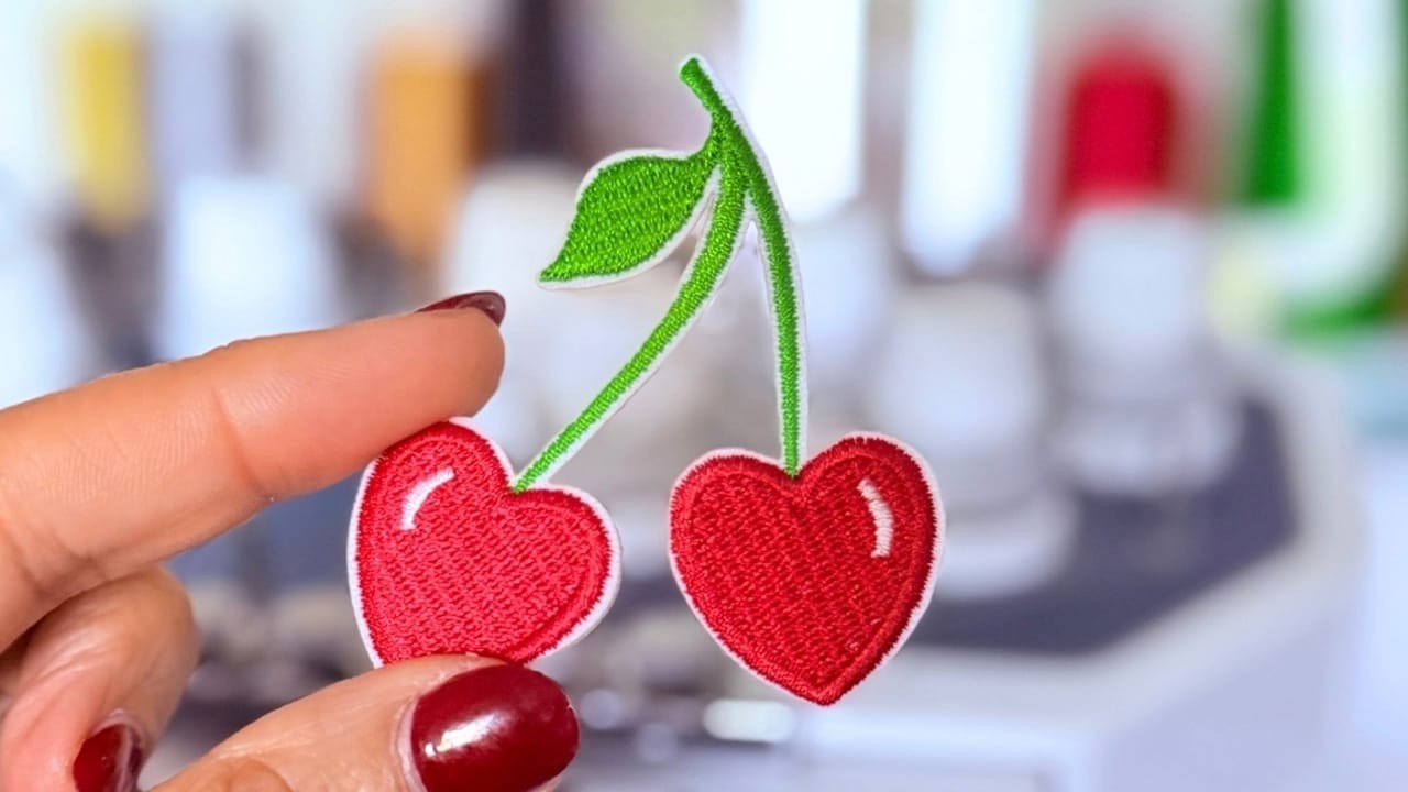 Patch brodé thermocollant cerises en forme de cœur, rouge vif, tenu à la main devant une machine à coudre – parfait pour personnaliser vêtements ou accessoires.