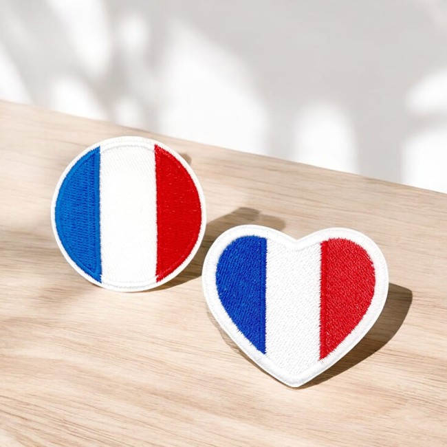 Duo d’écussons drapeau France, cœur et rond, mis en scène sur un fond bois clair, baigné de lumière naturelle.