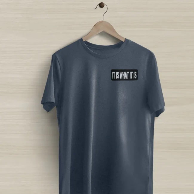 Écusson It Is What It Is sur un tee-shirt Patch It Is What It Is posé sur un tee-shirt bleu gris