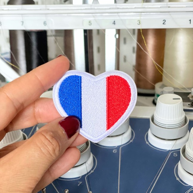 Patch cœur France brodé, tenu entre les doigts devant une machine à broder, montrant la fabrication artisanale et le soin du détail.