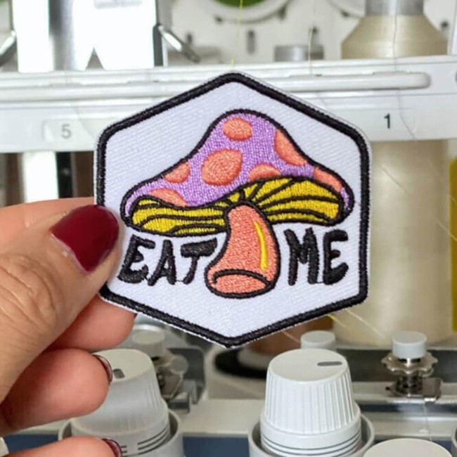 Ecusson psychédélique EAT ME avec bordure noire tenue dans ma main devant ma machine à broder patch brodé coloré fun et rétro