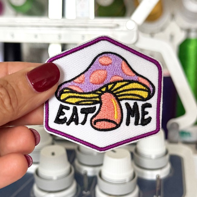 Ecusson psychédélique EAT ME avec bordure violette tenue dans ma main devant ma machine à broder patch brodé coloré fun et rétro