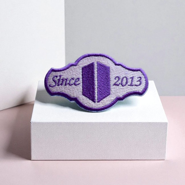 Patch ARMY BTS brodé – présentation sur socle pastel Patch ARMY BTS violet posé sur cube blanc et fond rose
