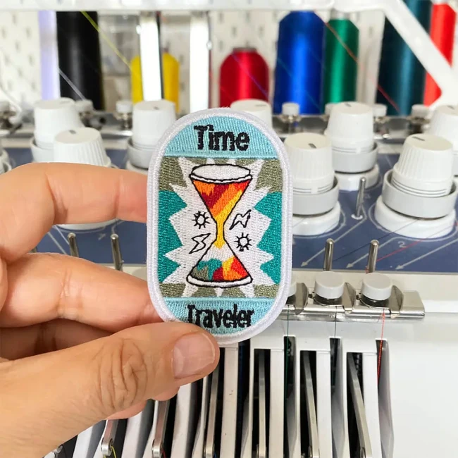 Écusson brodé photographié dans mon atelier de broderie Patch brodé Time Traveler sablier rétro tenu devant une machine à broder