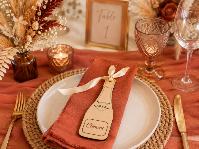 Porte-nom mariage en bois personnalisé en forme de bouteille de champagne présenté sur une table de mariage terracotta avec ruban satin
